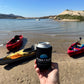 A hand holding up a can with a Floatsup coozie on it with Kayaks and a lake in the background. 